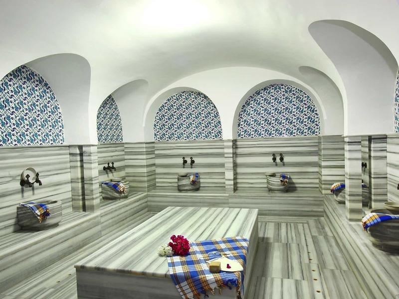 Elegant tiled spa room with arches, water taps, and benches, featuring a towel and flower on a table.