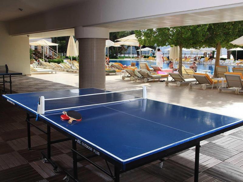 Blue ping pong table with paddles and ball near poolside lounge chairs under umbrellas.