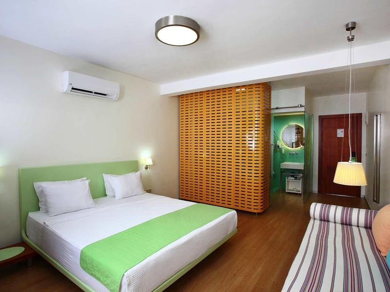 Modern hotel room with double bed, green accents, sofa, wooden floor, and air conditioning.