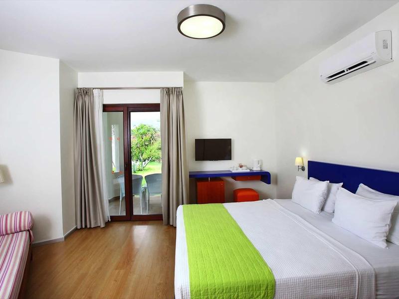Bright hotel room with double bed, green bed runner, and balcony door overlooking greenery.