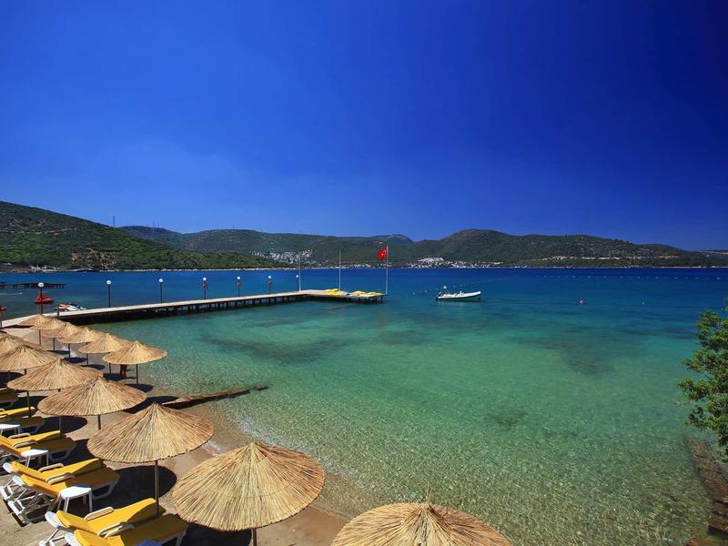Clear blue sea, thatched umbrellas, lounge chairs, a wooden pier, and green hills in the background.