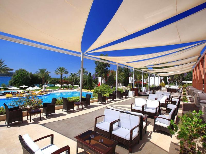 Outdoor seating area by a pool with shaded white cushioned chairs and palm trees under blue sky.