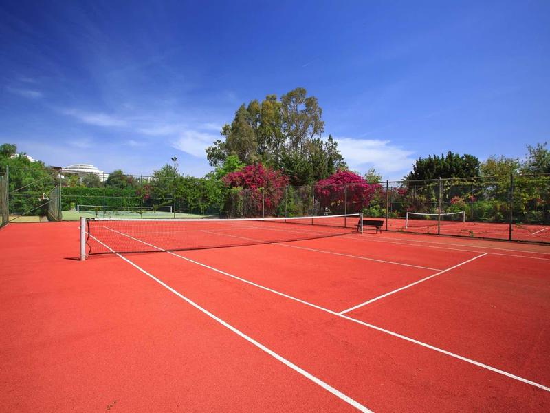 Bright red tennis court with net and green foliage under a clear blue sky.