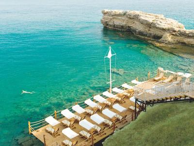 Eine Terrasse mit weißen Liegestühlen am klaren türkisen Meer neben Felsen und grünem Gras.
