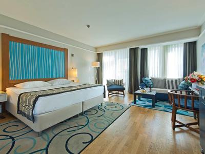 Licht ingericht hotelkamer met tweepersoonsbed, fauteuils en bureau.