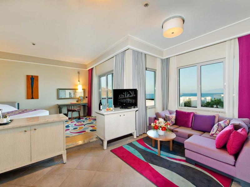 Modern hotelkamer met slaap- en woonruimte met paarse bank en groot raam.