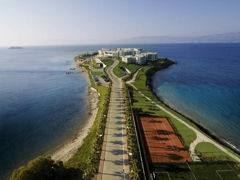 Lange landstrook met weg, tennisbanen en gebouwen aan zee onder een heldere hemel.