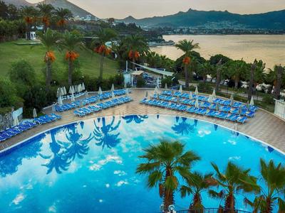 Großer blauer Pool mit Palmen und Liegestühlen, grüner Park und Berge im Hintergrund bei Sonnenuntergang.