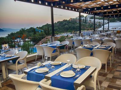 Terrasse eines Restaurants mit blauen Tischdecken und Blick auf Meer und Hügel bei Sonnenuntergang.