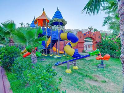 Bunter Spielplatz mit Rutsche und Wippe umgeben von Palmen und grünen Büschen unter blauem Himmel.