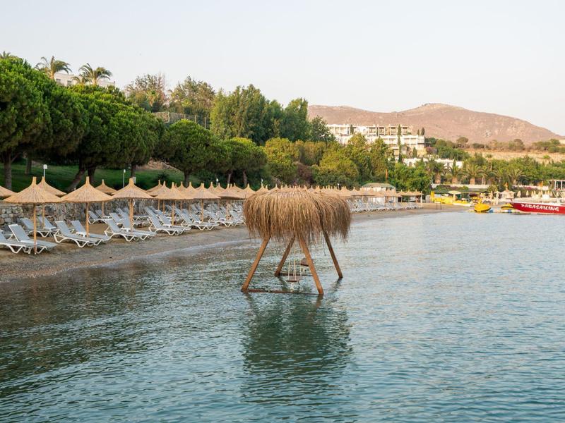 Strand mit Liegestühlen, Bäumen am Ufer und einer Holzskulptur im Wasser vor Bergen.
