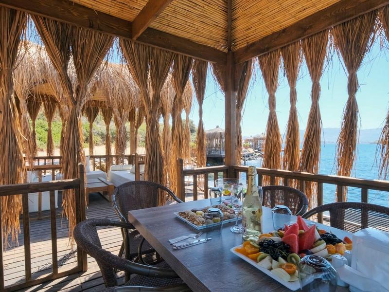 Überdachte Holzterrasse am Wasser mit gedecktem Tisch, Stühlen und Vorhängen aus getrocknetem Blattwerk.