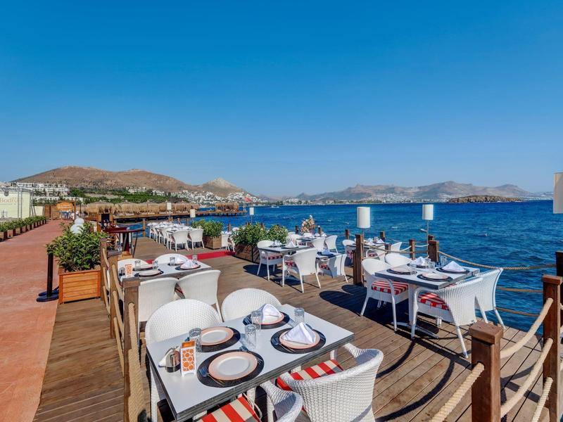 Terrassenrestaurant am Meer mit weißen Tischen, Stühlen und klar blauem Himmel und Wasser.