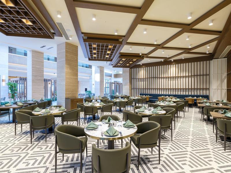 Przestronna jadalnia z okrągłymi stołami, zielonymi krzesłami i nowoczesnym wystrojem.