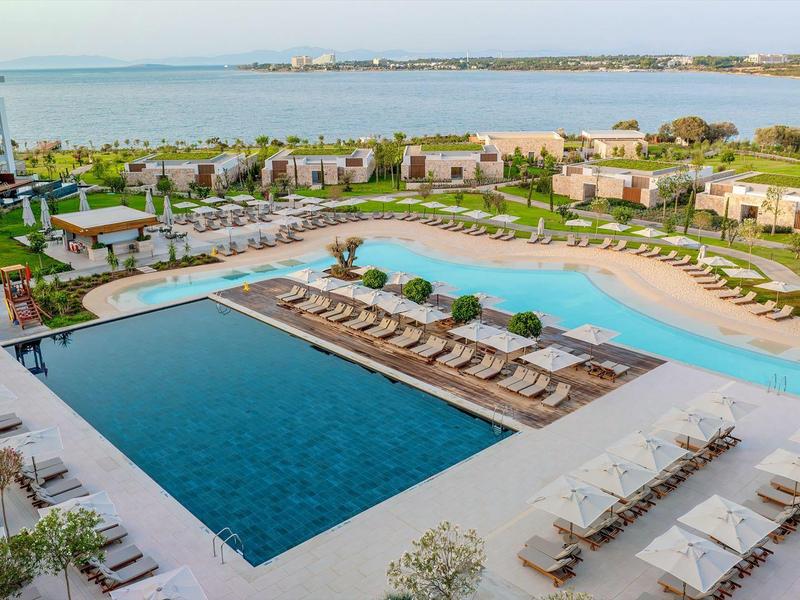 Duży basen hotelowy z leżakami i widokiem na morze w słonecznym otoczeniu.