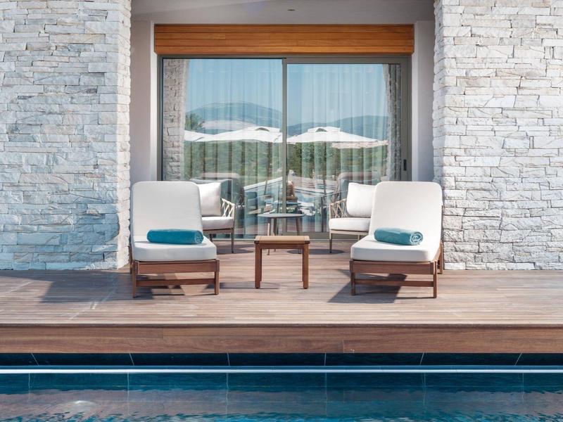 Dwa fotele z niebieskimi poduszkami na drewnianym tarasie przy basenie przed nowoczesnym domem z kamienia.