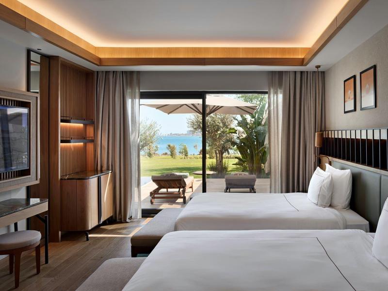 Nowoczesny pokój hotelowy z dwoma łóżkami, telewizorem, krzesłem i balkonem z widokiem na ogród i morze.