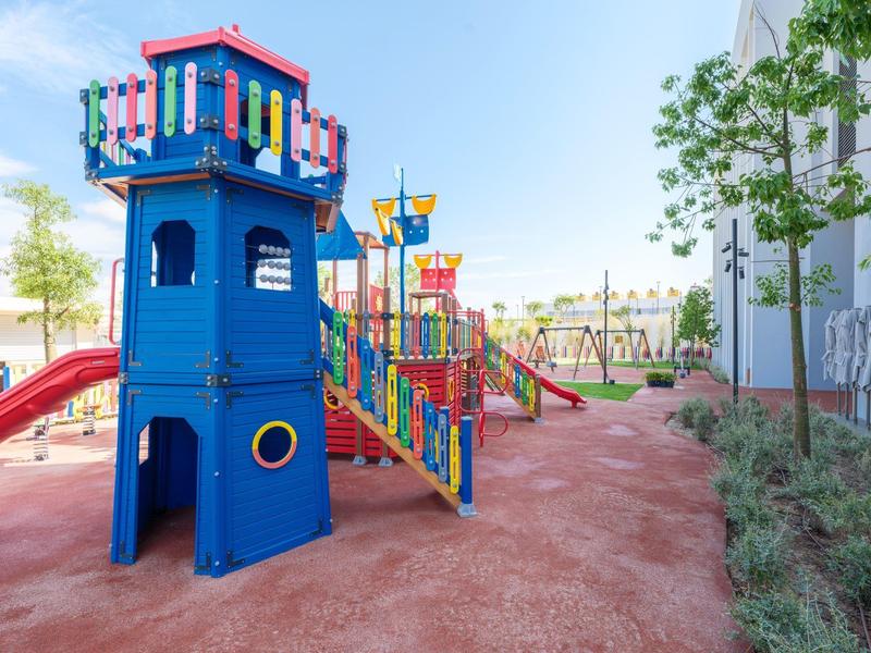 Kolorowy plac zabaw dla dzieci z zjeżdżalniami obok budynku i drzew.