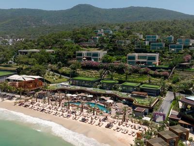 Luxuriöses Resort mit Pool, Sonnenschirmen und Liegestühlen am Strand vor bewaldeten Hügeln.