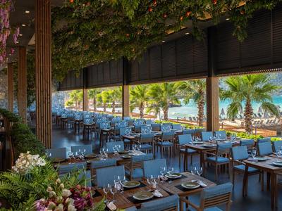 Offenes Restaurant mit Holztischen und Stühlen, Blick auf Palmen und Meer.