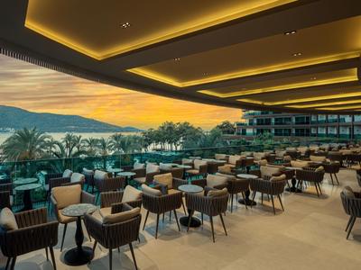 Moderner Hotelterrassenbereich mit vielen Tischen und Stühlen, Blick auf Palmen und Meer bei Sonnenuntergang.