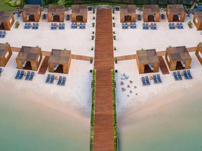 Ein Holzsteg führt zu einzelnen Strandhütten mit Liegestühlen am weißen Sandstrand und türkisem Wasser.