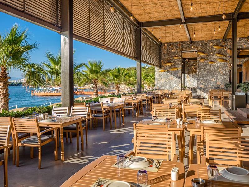 Otwarta restauracja z drewnianymi stołami i krzesłami, widokiem na palmy i morze pod błękitnym niebem.