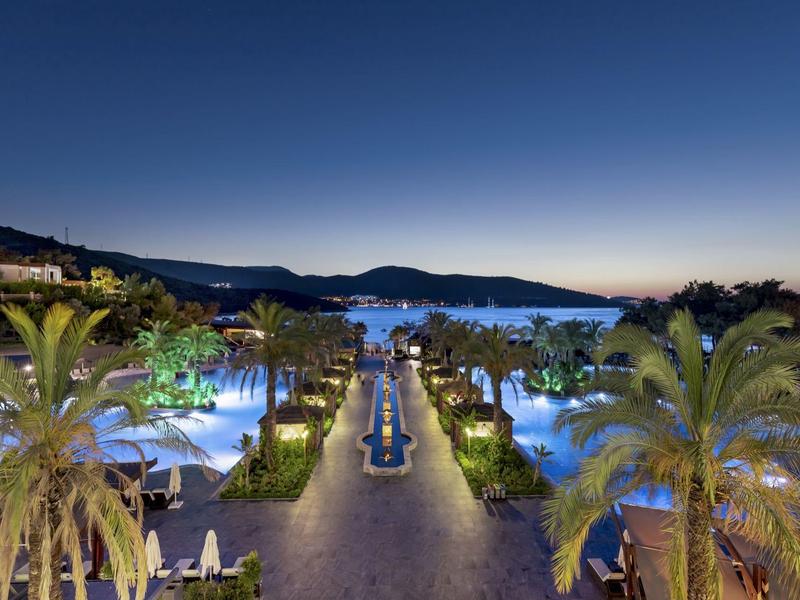 Nocny widok hotelu z oświetlonymi basenami i palmami wzdłuż ścieżki do morza.