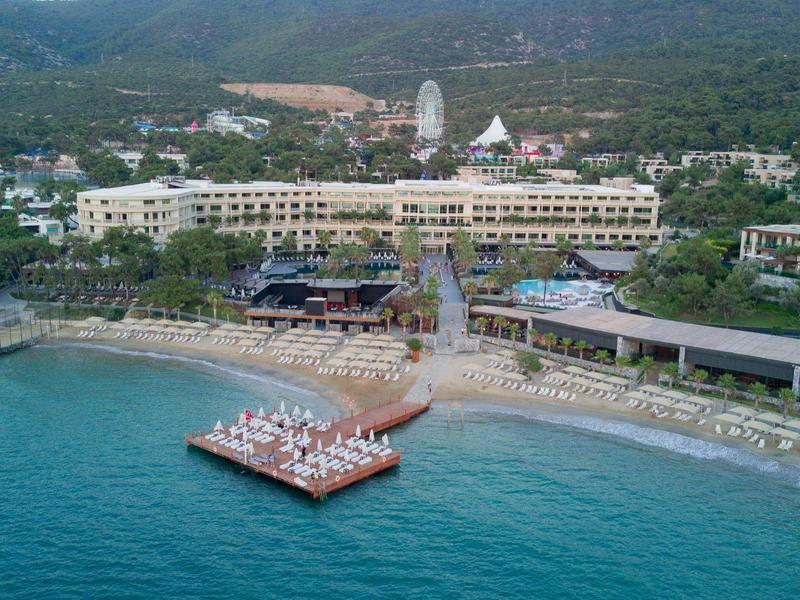 Luksusowy hotel przy plaży z basenem, leżakami i spokojnym morzem na górskim tle.