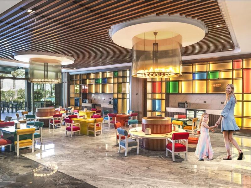 Nowoczesna, kolorowa kawiarnia z oświetleniem sufitowym, drewnianymi meblami i matką z dzieckiem w jasnym pomieszczeniu.