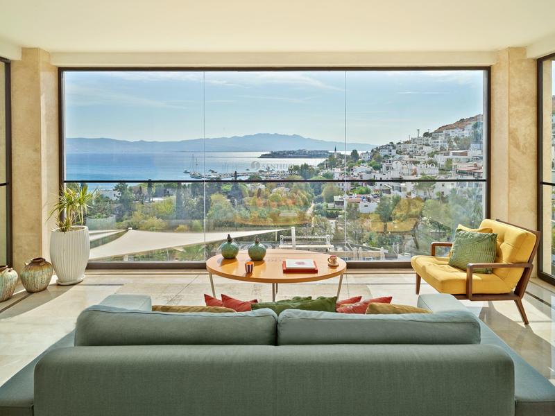 Jasny pokój z sofą, fotelami i dużym oknem z widokiem na morze i nadmorskie miasto.