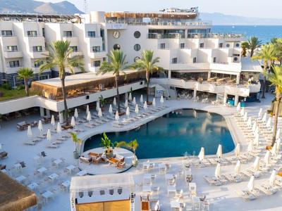 Hotel with large pool area, sun loungers, palm trees, and sea view in the background.