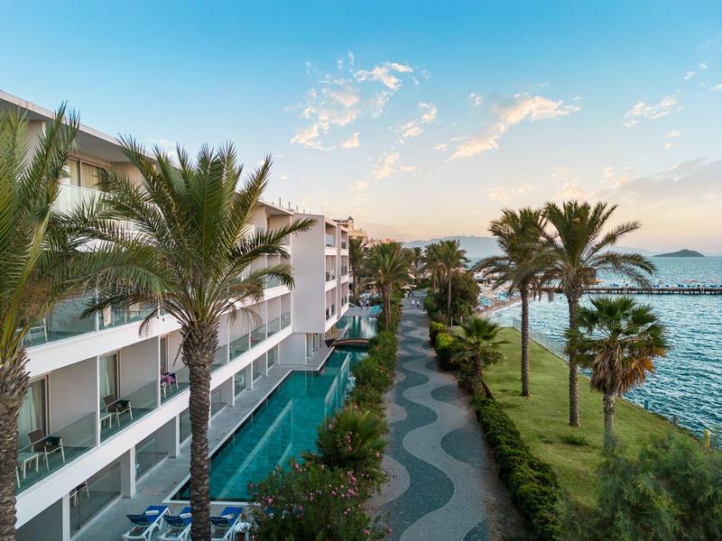 Hotel with palm trees, pool, and sea view under clear evening sky.
