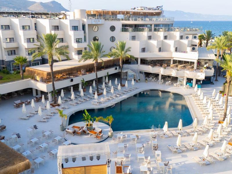 Hotel with large pool area, sun loungers, palm trees, and sea view in the background.