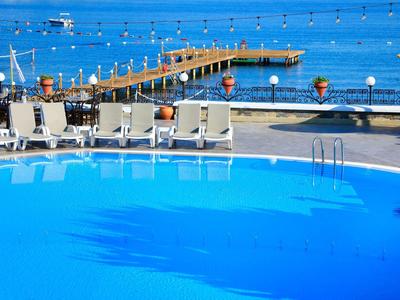 Área de piscina vacía con tumbonas y vista a un muelle sobre el mar azul bajo cielo despejado.
