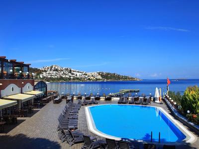 Pohled na hotelový bazén s lehátky a mořem v pozadí za jasné oblohy.