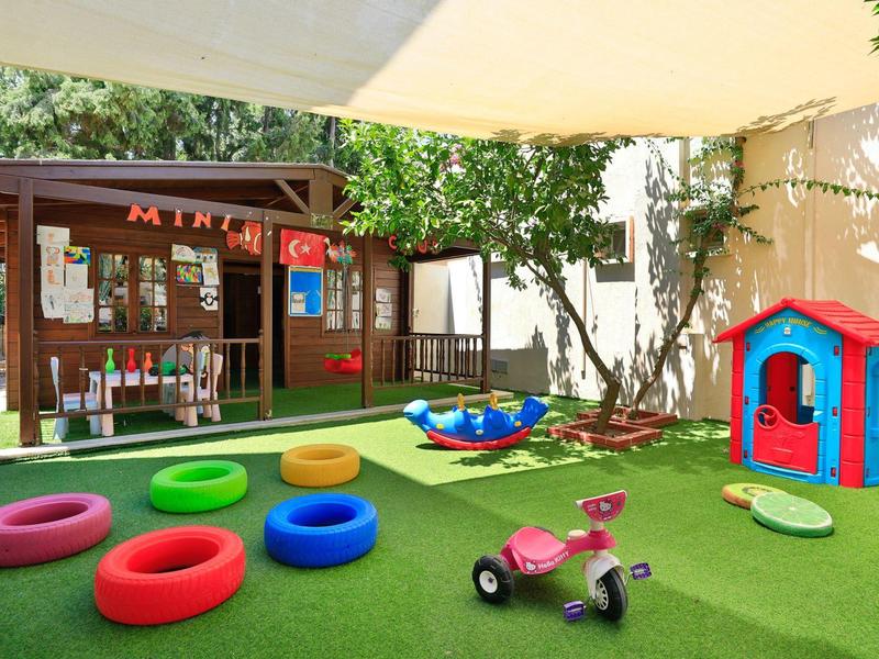 Área de juegos infantiles con juguetes, aros de salto y una casita pequeña sobre césped artificial bajo sombra.