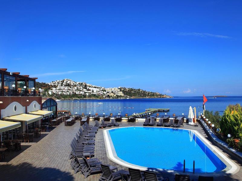 Piscina del hotel con tumbonas, vista al mar y la ciudad costera bajo un cielo azul claro.