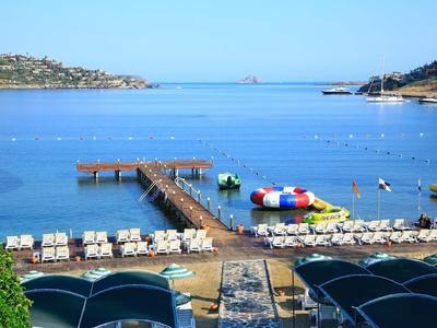Strand mit Holzsteg, Liegen und Wasserspielgeräten vor ruhigem blauem Meer bei klarem Himmel.