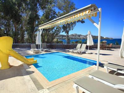 Piscine avec pare-soleil et toboggan jaune à côté de transats, vue sur la mer et les collines.