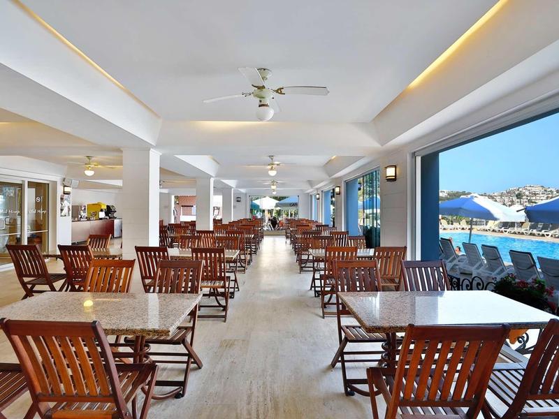Grande salle à manger lumineuse avec tables en bois et vue sur la piscine et la mer.