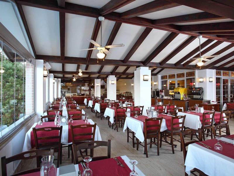 Restaurant avec nappes rouges, plafond à poutres en bois et grandes fenêtres donnant sur un jardin.