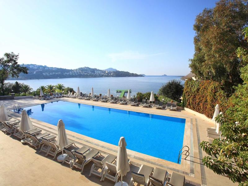 Une grande piscine avec des chaises longues et des parasols à côté d'une eau calme et des collines boisées en arrière-plan.