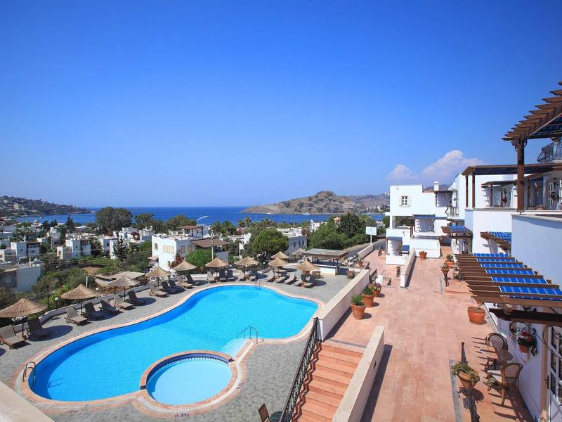 Schwimmbad mit klarem Wasser, umgeben von Sonnenschirmen, weißer Häuser und blauem Himmel.