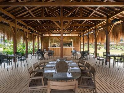 Restaurant en plein air avec tables en bois sous un toit en chaume dans un cadre naturel.