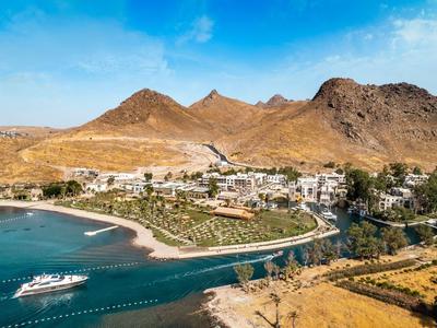Vue côtière d'une baie avec yacht, village et montagnes arides sous ciel bleu.
