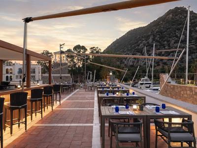 Terrasse de restaurant en bord de mer avec vues sur bateaux et collines au coucher du soleil.