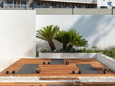 Outdoor-Yogabereich mit Holzplattformen, Matten und kleinen Gewichten vor weißer Wand und Pflanzen.