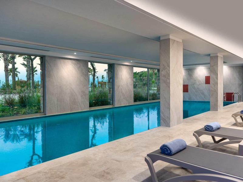 Piscine intérieure avec chaises longues et fenêtres donnant sur des palmiers.