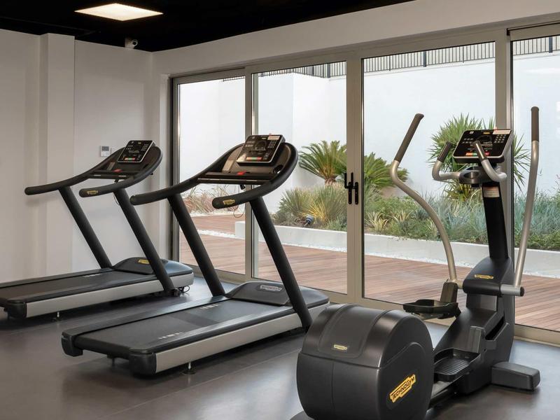 Salle de sport lumineuse avec tapis de course et vélo elliptique près de grandes fenêtres.